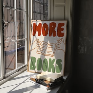 Peut inclure: Une affiche encadr&eacute;e avec le texte "MORE BOOKS" en lettres orange et vertes. L'affiche pr&eacute;sente une image de dessin anim&eacute; de deux mains tenant un livre ouvert. L'affiche est pos&eacute;e sur un rebord de fen&ecirc;tre avec une pile de livres en dessous.