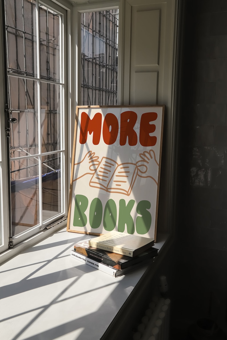 Peut inclure: Une affiche encadr&eacute;e avec le texte "MORE BOOKS" en lettres orange et vertes. L'affiche pr&eacute;sente une image de dessin anim&eacute; de deux mains tenant un livre ouvert. L'affiche est pos&eacute;e sur un rebord de fen&ecirc;tre avec une pile de livres en dessous.