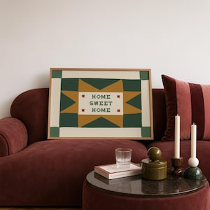 Puede incluir: Una l&aacute;mina de arte rectangular enmarcada con un dise&ntilde;o de estrella estilizado en amarillo mostaza y verde oscuro sobre un fondo crema. El centro de la estrella contiene el texto "HOME SWEET HOME" en letras verde oscuro, flanqueado por cuatro peque&ntilde;os iconos de estrellas rojas.