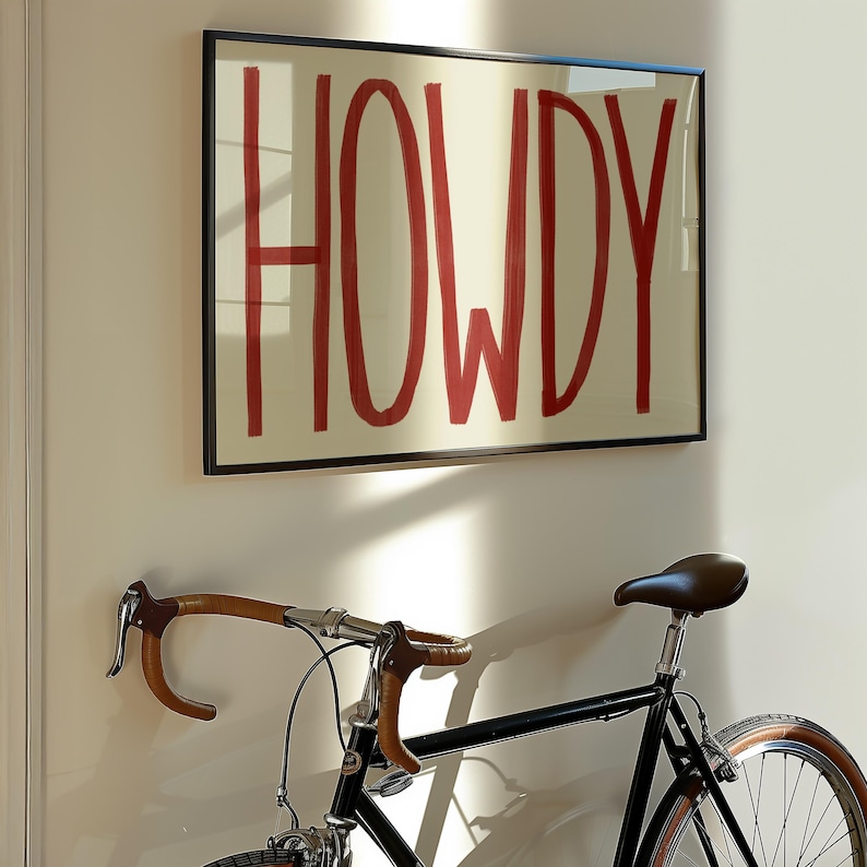 Puede incluir: Una impresi&oacute;n enmarcada con la palabra "HOWDY" escrita en rojo sobre un fondo crema. Una bicicleta negra con manillar marr&oacute;n est&aacute; en primer plano.