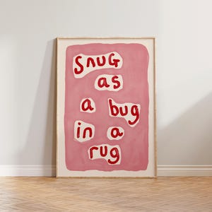 May include: A framed art print with the phrase "Snug as a bug in a rug" in red letters on a pink background. The artwork has a simple, hand-painted style and is displayed in a light-colored wooden frame.