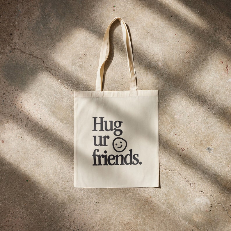 May include: A white canvas tote bag with black text that reads "Hug ur friends." and a black smiley face.