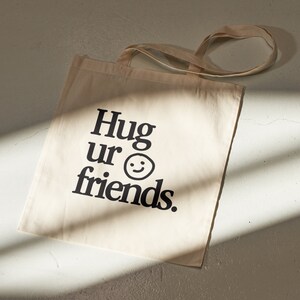 May include: A beige canvas tote bag with black text that reads "Hug ur friends." and a simple smiley face.