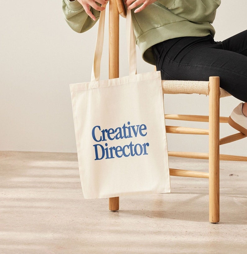 May include: A white canvas tote bag with blue text that reads "Creative Director".