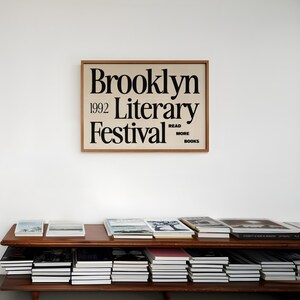 May include: A wooden-framed poster for the 1992 Brooklyn Literary Festival, with black text on a cream background, hangs above a shelf of books. The poster includes the words "Read More Books". The shelf displays stacks of books.
