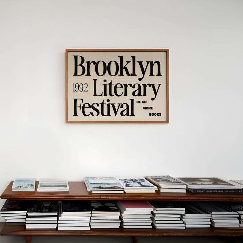 May include: A wooden-framed poster for the 1992 Brooklyn Literary Festival, with black text on a cream background, hangs above a shelf of books. The poster includes the words "Read More Books". The shelf displays stacks of books.
