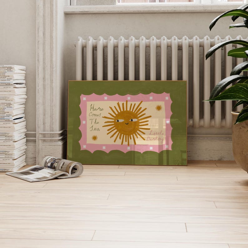 Puede incluir: Una impresi&oacute;n art&iacute;stica enmarcada con una ilustraci&oacute;n de un sol sonriente y el texto "Here Comes The Sun, Little Darling". La obra de arte tiene un borde verde, un borde interior rosa festoneado y un fondo crema.