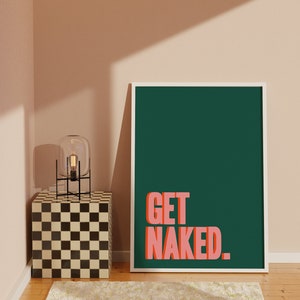 May include: A rectangular wall art print in a white frame featuring a dark green background with the text GET NAKED in bold, salmon-pink, block-style lettering. The print is positioned upright on a wooden floor next to a checkered cube side table.