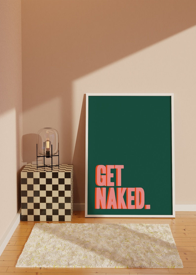 May include: A rectangular wall art print in a white frame featuring a dark green background with the text GET NAKED in bold, salmon-pink, block-style lettering. The print is positioned upright on a wooden floor next to a checkered cube side table.