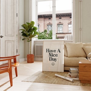 May include: A beige couch with a throw pillow sits in a living room with a large window. A framed print with the text "Have A Nice Day" and a smiley face hangs on the wall above a wooden coffee table. A potted plant sits in the corner of the room.