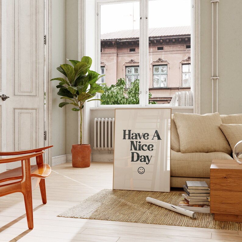 May include: A beige couch with a throw pillow sits in a living room with a large window. A framed print with the text "Have A Nice Day" and a smiley face hangs on the wall above a wooden coffee table. A potted plant sits in the corner of the room.