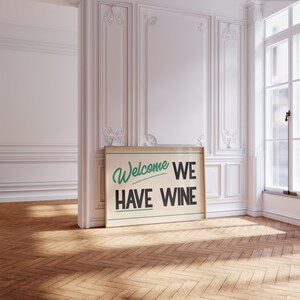 May include: A framed poster with a white background and black text that reads "Welcome We Have Wine". The text is in a retro style font with a green underline. The poster is hanging on a white wall in a room with white moldings.