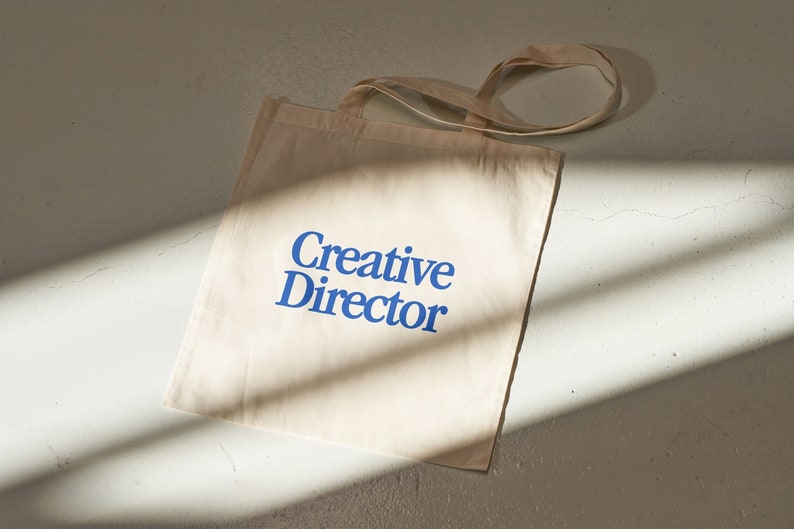 May include: A beige canvas tote bag with blue text that reads "Creative Director".