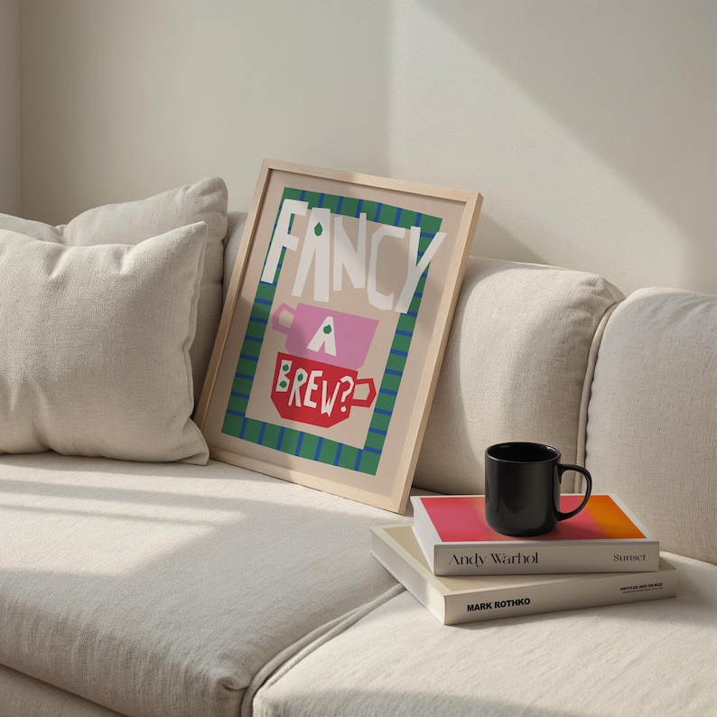 Op de afbeelding: Een ingelijst print met een kleurrijk ontwerp met de tekst "Fancy a Brew?" met een groene, roze en rode theekopillustratie. De print is op een witte bank met een stapel boeken.
