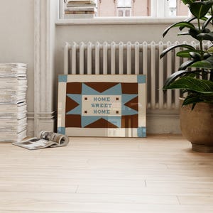 Puede incluir: Una impresi&oacute;n de arte mural rectangular con un fondo marr&oacute;n y una gran estrella de ocho puntas de color azul claro en el centro. El texto "HOME SWEET HOME" est&aacute; impreso en letras de bloque azul claro dentro de la estrella, flanqueado por cuatro peque&ntilde;as estrellas marrones.