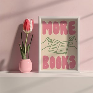 Peut inclure: Une impression encadr&eacute;e blanche avec le texte "MORE BOOKS" en lettres roses. L'impression pr&eacute;sente une illustration verte d'un livre ouvert avec des mains de chaque c&ocirc;t&eacute;. Une fleur rose dans un vase rose se trouve devant l'impression.