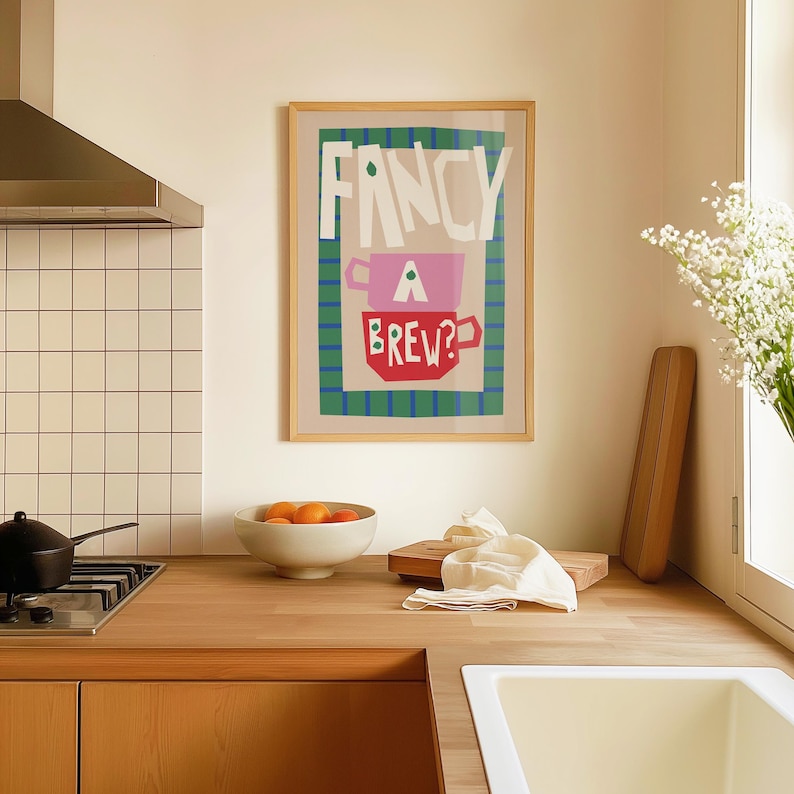 Op de afbeelding: Een ingelijste poster met de tekst "FANCY A BREW?" in kleurrijke, gestileerde letters. De poster toont twee gestapelde theekopjes, een roze en een rode, met groene accenten.