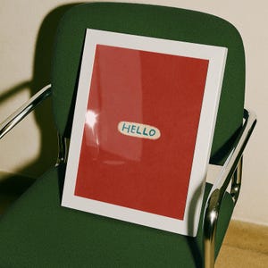 Puede incluir: Una obra de arte enmarcada con un borde blanco y un fondo rojo. La obra presenta una peque&ntilde;a pegatina ovalada con la palabra "HELLO" en letras azules. El marco descansa sobre una silla verde con reposabrazos plateados.