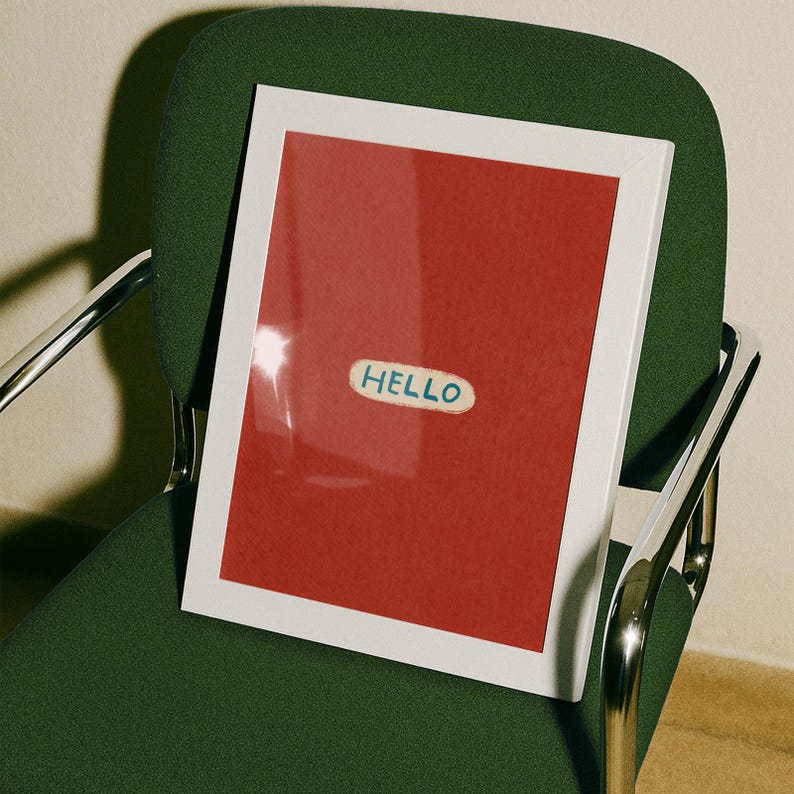 Puede incluir: Una obra de arte enmarcada con un borde blanco y un fondo rojo. La obra presenta una peque&ntilde;a pegatina ovalada con la palabra "HELLO" en letras azules. El marco descansa sobre una silla verde con reposabrazos plateados.