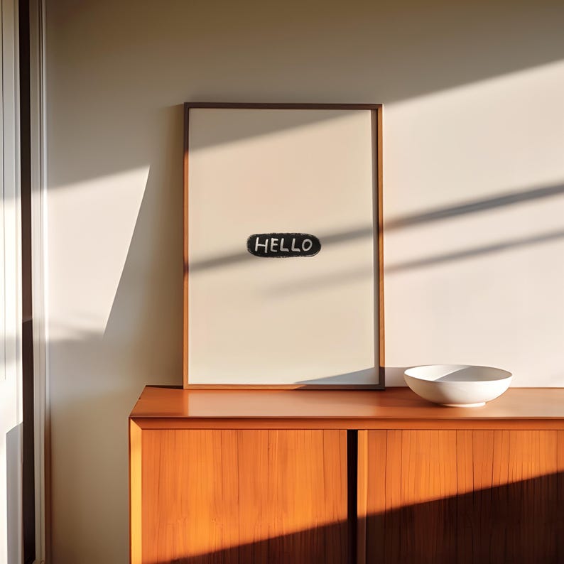 Puede incluir: Una impresi&oacute;n enmarcada con la palabra "HELLO" en un &oacute;valo negro. La obra de arte se exhibe en un mueble de madera, con un cuenco blanco. La habitaci&oacute;n est&aacute; iluminada por luz natural, creando sombras.