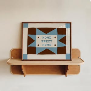 Puede incluir: Una l&aacute;mina de arte mural rectangular en un marco de madera oscura con un patr&oacute;n geom&eacute;trico de estrellas en marr&oacute;n y azul claro sobre un fondo crema. El centro del dise&ntilde;o muestra el texto HOME SWEET HOME en letras azules con estrellas marrones.
