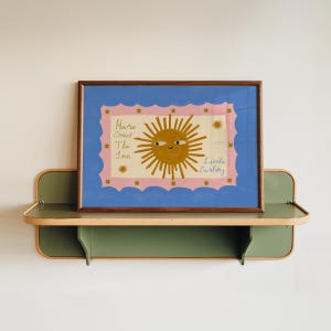 Puede incluir: Una impresi&oacute;n art&iacute;stica enmarcada con una ilustraci&oacute;n de un sol sonriente y el texto "Here Comes The Sun Little Darling". La obra de arte tiene un borde azul y un fondo rosa. El marco es marr&oacute;n y la impresi&oacute;n se muestra en una estanter&iacute;a verde y beige.