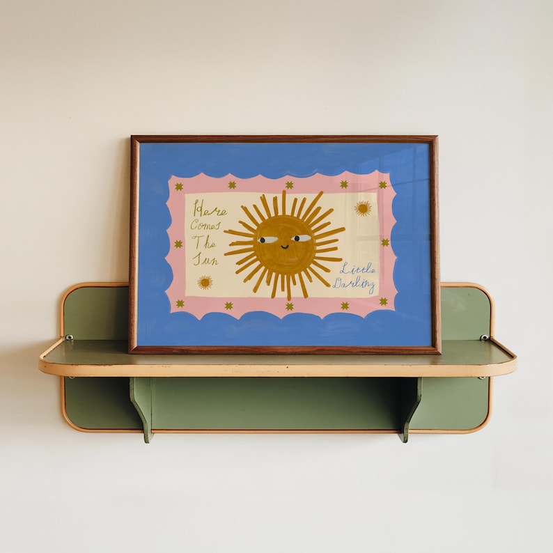 Puede incluir: Una impresi&oacute;n art&iacute;stica enmarcada con una ilustraci&oacute;n de un sol sonriente y el texto "Here Comes The Sun Little Darling". La obra de arte tiene un borde azul y un fondo rosa. El marco es marr&oacute;n y la impresi&oacute;n se muestra en una estanter&iacute;a verde y beige.