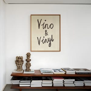 Puede incluir: Una obra de arte enmarcada con las palabras "Vino & Vinyl" en escritura negra cuelga sobre una estanter&iacute;a de madera. La estanter&iacute;a muestra pilas de libros y dos peque&ntilde;as esculturas. La habitaci&oacute;n tiene un dise&ntilde;o minimalista con paredes blancas.