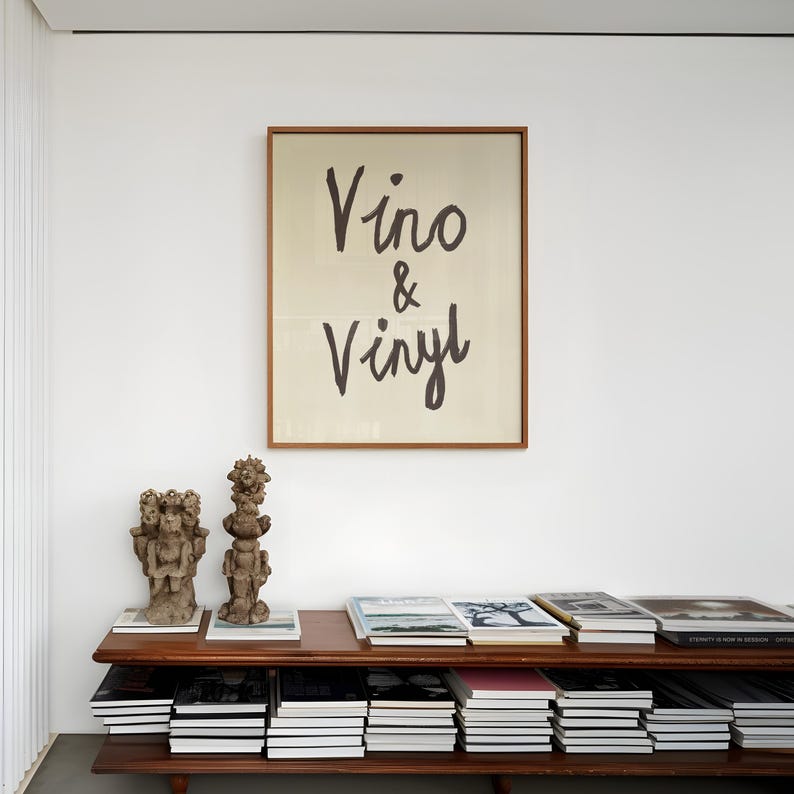 Puede incluir: Una obra de arte enmarcada con las palabras "Vino & Vinyl" en escritura negra cuelga sobre una estanter&iacute;a de madera. La estanter&iacute;a muestra pilas de libros y dos peque&ntilde;as esculturas. La habitaci&oacute;n tiene un dise&ntilde;o minimalista con paredes blancas.