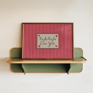 K&ouml;nnte beinhalten: Ein gerahmter Druck mit einem rot-rosa gestreiften Hintergrund und dem Text "Night Night Love You". Der Druck wird auf einem gr&uuml;nen und holzfarbenen Regal pr&auml;sentiert, was eine charmante, Vintage-&Auml;sthetik erzeugt.