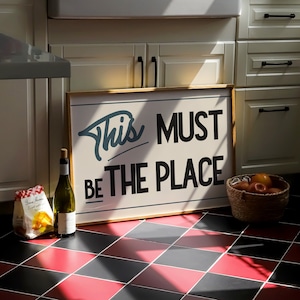 May include: A wooden sign with white text that reads "This must be the place". The sign is hanging on a wall with white cabinets and a checkered black and red floor.