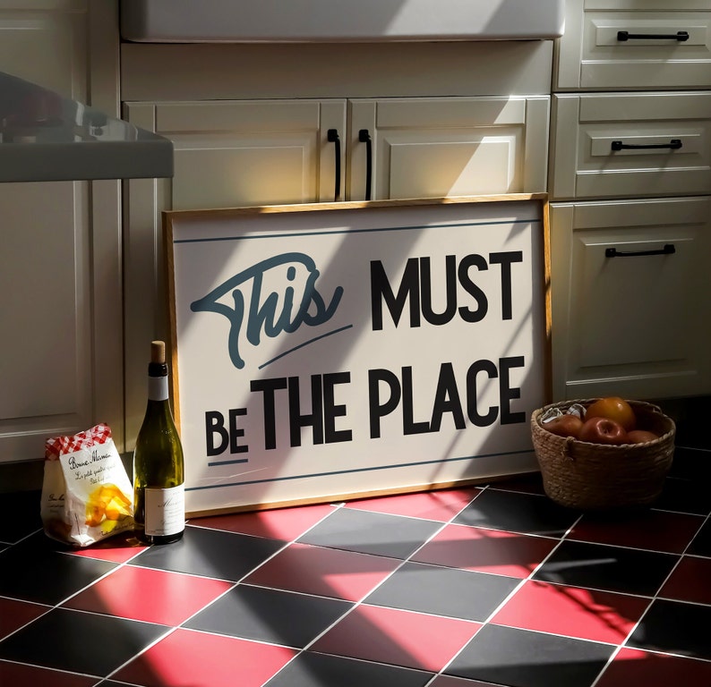May include: A wooden sign with white text that reads "This must be the place". The sign is hanging on a wall with white cabinets and a checkered black and red floor.