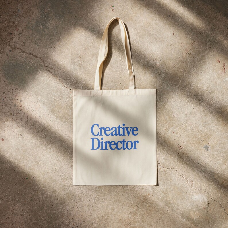 May include: A white canvas tote bag with blue text that reads "Creative Director".