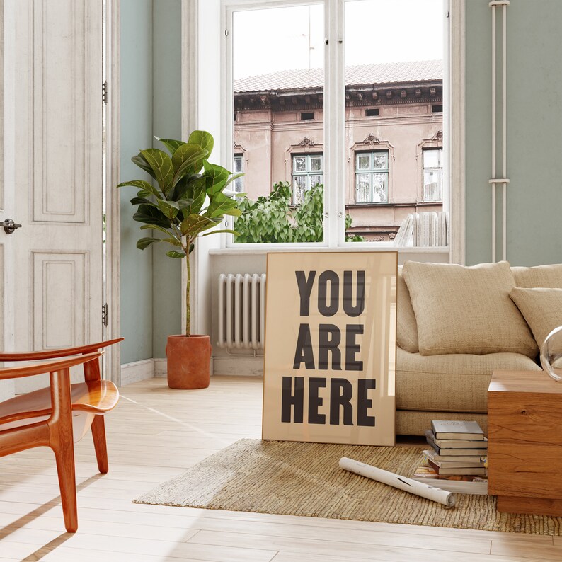 Peut inclure: Un canap&eacute; beige avec un coussin est plac&eacute; dans un salon avec une grande fen&ecirc;tre. Un poster encadr&eacute; avec le texte "YOU ARE HERE" est appuy&eacute; contre le canap&eacute;. Une table basse en bois avec des livres et un vase se trouve devant le canap&eacute;.