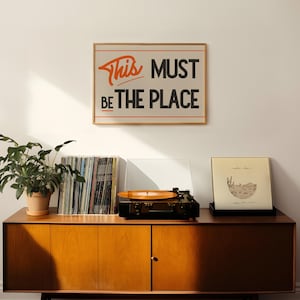 May include: A framed print with the text "This Must Be The Place" in orange and black lettering on a beige background. The print is hanging on a white wall above a wooden cabinet with a record player, a stack of vinyl records, and a potted plant.