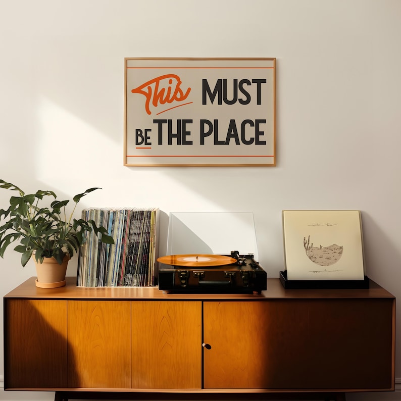 May include: A framed print with the text "This Must Be The Place" in orange and black lettering on a beige background. The print is hanging on a white wall above a wooden cabinet with a record player, a stack of vinyl records, and a potted plant.