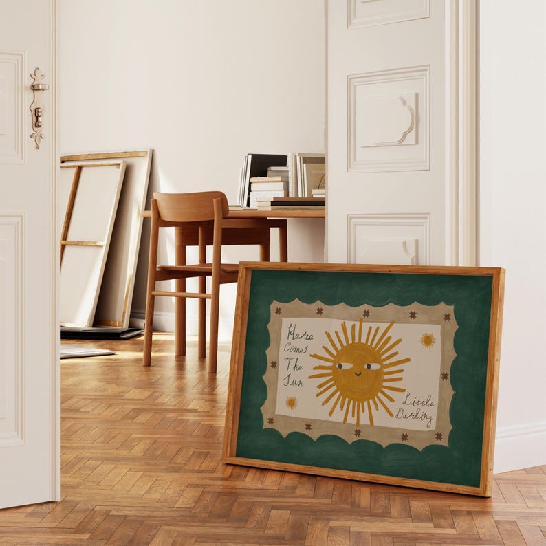 Puede incluir: Una impresi&oacute;n art&iacute;stica enmarcada con una ilustraci&oacute;n de un sol sonriente y el texto "Here Comes The Sun" y "Little Darling". La obra de arte tiene un borde verde festoneado y un marco de madera. El fondo muestra una habitaci&oacute;n con suelo de madera y un escritorio.