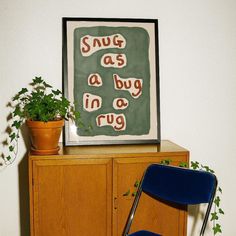 May include: A framed art print with the phrase "snug as a bug in a rug" in white letters on a green background. The print is displayed on a wooden cabinet, with a potted plant and a blue chair in the foreground.