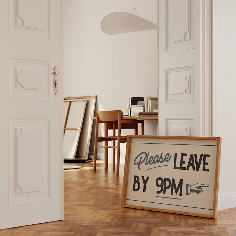 May include: A framed sign with the text "Please LEAVE BY 9PM" in a vintage style. The sign is in a room with a wooden floor, a desk, and a chair. The sign has a wooden frame and a beige background.