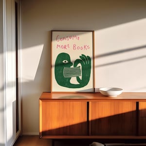 May include: A framed illustration of a green figure with a mouth covered by a striped mask. The text "CONSUME MORE BOOKS" is written in pink above the figure.
