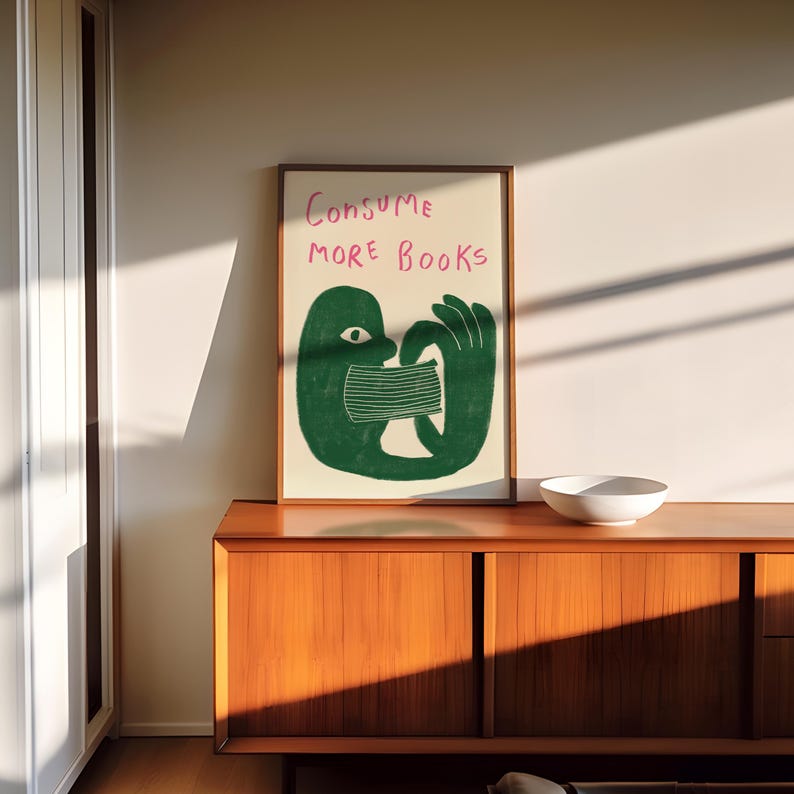 May include: A framed illustration of a green figure with a mouth covered by a striped mask. The text "CONSUME MORE BOOKS" is written in pink above the figure.