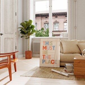 Puede incluir: Una impresi&oacute;n enmarcada con el texto "This Must Be The Place" en letras de colores sobre un fondo claro. La impresi&oacute;n est&aacute; colgada en una pared en una sala de estar con un sof&aacute;, una silla, una planta y una ventana.
