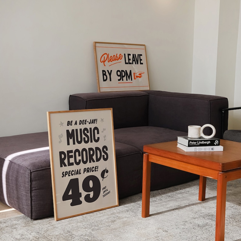 May include: Two framed posters with text, "Please Leave by 9PM" and "Music Records Special Price 49". A dark gray sectional sofa and a wooden coffee table with books and mugs are also visible.