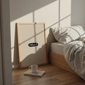 Puede incluir: Una impresi&oacute;n art&iacute;stica enmarcada con la palabra "HELLO" en una burbuja negra. La impresi&oacute;n est&aacute; apoyada contra una pared junto a una cama con una colcha blanca. Una taza blanca y un libro est&aacute;n en el suelo frente a la impresi&oacute;n.