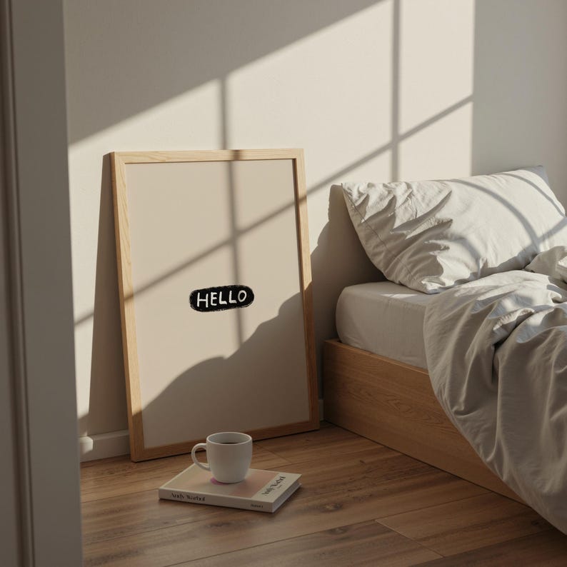 Puede incluir: Una impresi&oacute;n art&iacute;stica enmarcada con la palabra "HELLO" en una burbuja negra. La impresi&oacute;n est&aacute; apoyada contra una pared junto a una cama con una colcha blanca. Una taza blanca y un libro est&aacute;n en el suelo frente a la impresi&oacute;n.