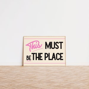 May include: A framed print with a white background and black text that reads "This must be the place". The word "This" is written in pink cursive.