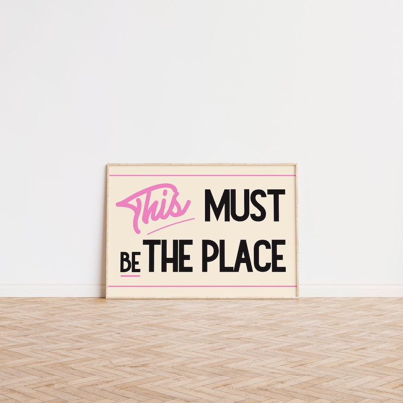 May include: A framed print with a white background and black text that reads "This must be the place". The word "This" is written in pink cursive.