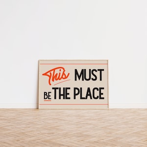 May include: A beige framed poster with the text "This Must Be The Place" in black and orange lettering.