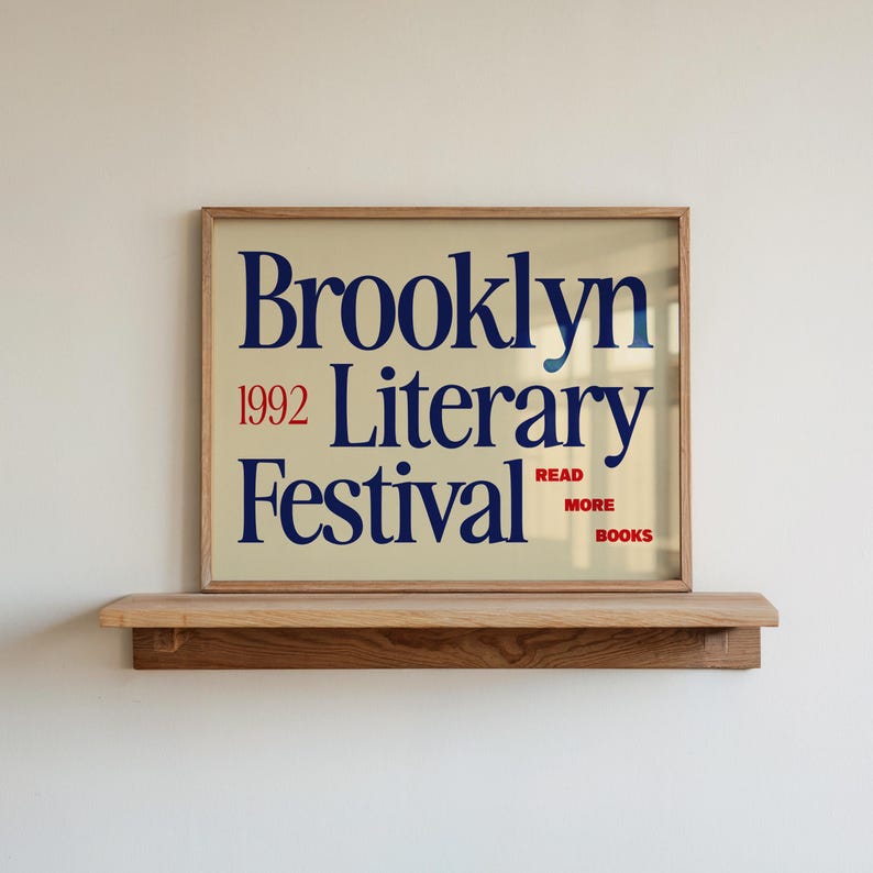 May include: A framed poster for the Brooklyn Literary Festival 1992. The poster has a beige background with navy blue text that reads "Brooklyn Literary Festival" and red text that reads "1992" and "Read More Books". The poster is displayed on a wooden shelf.