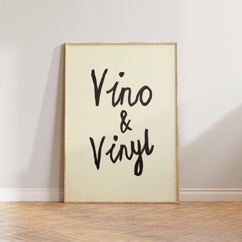 Puede incluir: Una impresi&oacute;n art&iacute;stica enmarcada con las palabras "Vino & Vinyl" en letras negras con pincel. El fondo es de color crema claro, y el marco es de tono madera clara. La impresi&oacute;n se muestra contra una pared blanca y un suelo de madera.